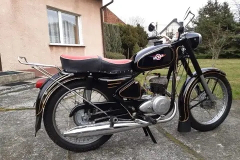 Czarny motocykl WSK z czerwoną kanapą i złotymi ozdobami, stojący na podjeździe.