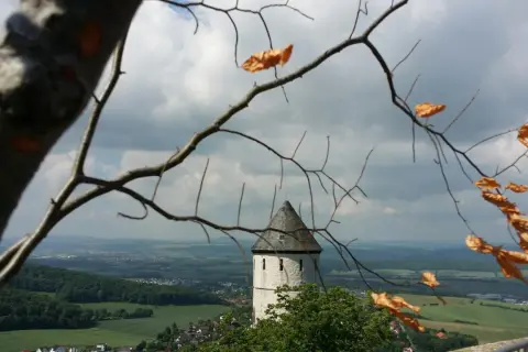 Blick durch kahle Äste auf die Burg Bielstein, ein weißer Turm mit grauem Dach ragt über grüne Hügel und eine Stadt.