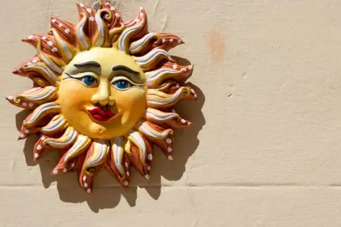 Sole decorativo in ceramica, un esempio di artigianato italiano, con volto sorridente e raggi colorati su muro beige.