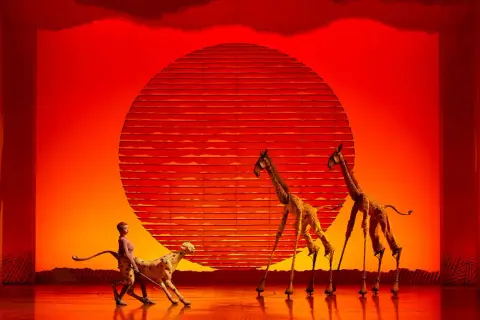 Escena de musicales en Zaragoza: un guepardo y dos jirafas en un escenario con un sol rojo.