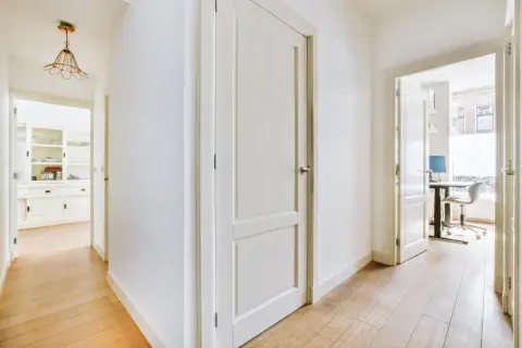Pasillo con puertas de interior blancas y suelo de madera. Se vislumbran una cocina y un despacho.