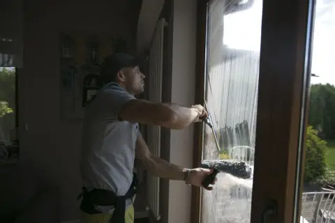 Mężczyzna w czapce z daszkiem pokazuje, jak myć okna ściągaczką, usuwając pianę z szyby.
