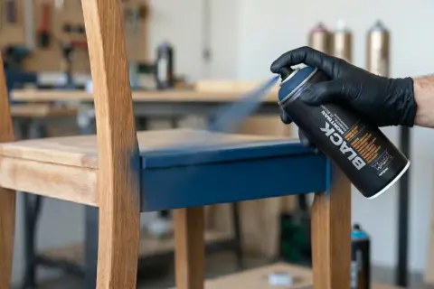 Main d'une personne portant un gant noir pulvérise de la peinture bleue sur un siège de chaise en bois, transformant un meuble mélaminé.