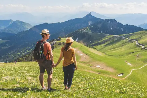 Couple en chapeau admirant un paysage de montagne verdoyant, parsemé de fleurs jaunes. Une rando Chartreuse insolite, avec des sentiers sinueux et des sommets bleutés à l'horizon.