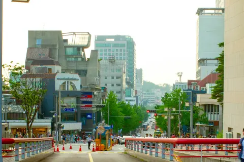 Tętniąca życiem ulica w **Hongdae**, pełna ludzi, sklepów i nowoczesnej architektury, z widokiem na wzgórza w tle.
