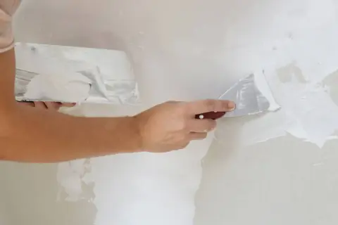Manos aplicando masilla blanca en una pared con espátulas. La mejor masilla para paredes deja un acabado liso y perfecto.