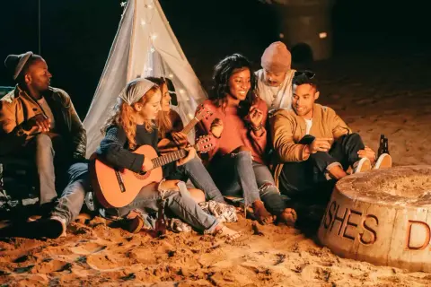 Freunde sitzen am Strand um ein Feuer, spielen Gitarre und singen Lagerfeuerlieder zum Mitsingen. Ein Zelt mit Lichterkette im Hintergrund.