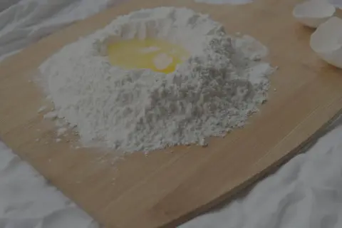 En hög mjöl med ett ägg i mitten, redo att blandas. Kanske lite för mycket bakpulver i receptet? Äggskal ligger bredvid.