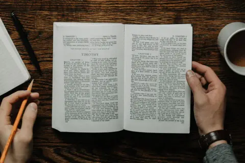 Dłonie trzymają otwartą księgę, jedną z apostolskich ksiąg Nowego Testamentu, obok leży notatnik i kubek kawy.