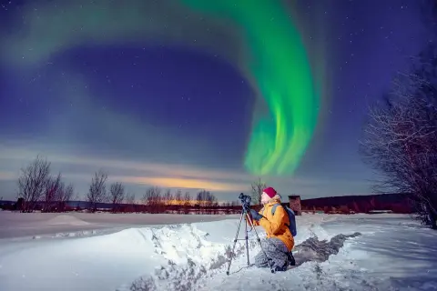 Fotograf uwiecznia zorzę polarną podczas zimowej wyprawy. Zielona poświata tańczy na nocnym niebie nad ośnieżonym krajobrazem.
