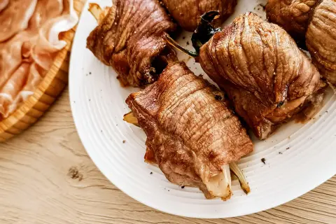 Involtini di carne succulenti, cotti alla perfezione, serviti su un piatto bianco. A lato, fette di prosciutto crudo in un cestino di legno.
