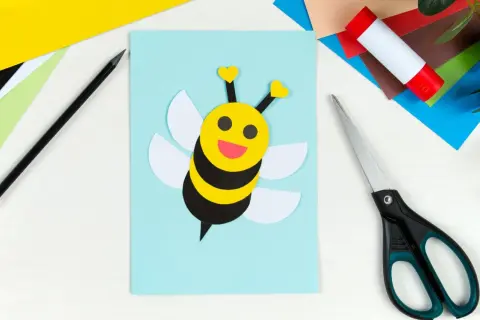Ein süßes Biene basteln-Projekt auf hellblauem Papier. Bunte Bastelmaterialien, Schere und Klebestift liegen bereit.