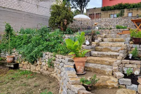 Ein Garten mit üppigen Tomatenpflanzen, begrenzt von einer niedrigen trockenmauer ohne fundament. Steintreppen führen zu einem Terrassenbereich.