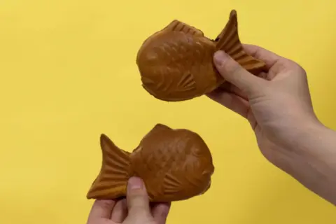 Zwei goldbraune Taiyaki, beliebte japanische Desserts in Fischform, werden von Händen auf gelbem Hintergrund gehalten.