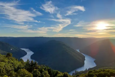 Un río serpentea entre montañas verdes, bajo un cielo azul con nubes finas. El sol se pone, tiñendo el horizonte de dorado. Parece el cabo do mundo.
