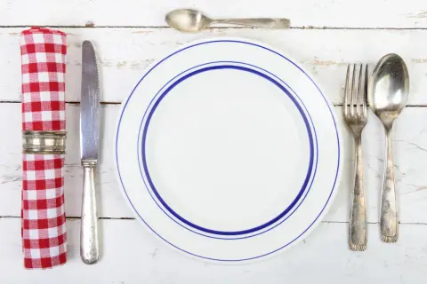 Une assiette blanche à double bordure bleue, une serviette à carreaux rouges et blancs, un couteau, une fourchette et deux cuillères. Qui a inventé les couverts ?