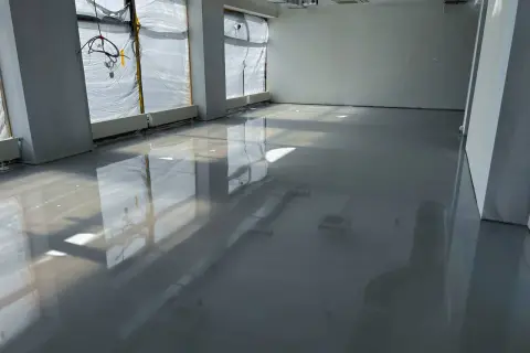 Lśniąca podłoga z żywicy w pomieszczeniu w budowie. Widać odbicia okien i instalacji.