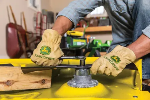 Gloves on, a person uses a socket wrench to remove a mower blade, preparing to sharpen it.