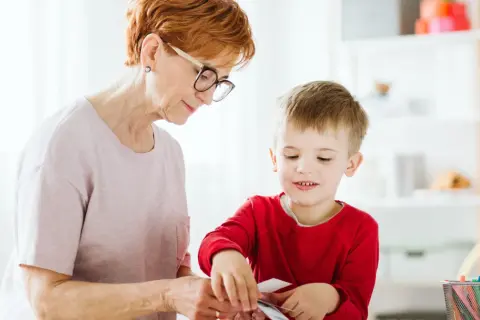 Babcia i wnuczek tworzą coś razem. Dziecko z zaciekawieniem przygląda się swoim ruchom, a jego manieryzmy ruchowe są widoczne w skupieniu.
