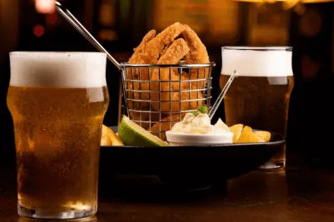 Due bicchieri di birra bionda ghiacciata accompagnano un cestino di anelli di cipolla fritti, patatine e una salsa cremosa.