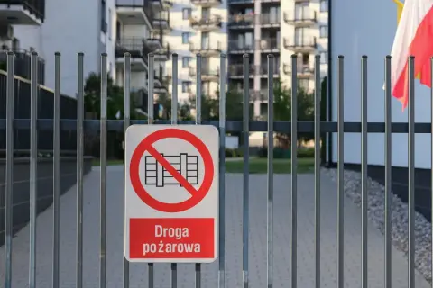 Znak zakazu wjazdu na drogę pożarową na metalowej bramie. W tle budynki mieszkalne i polska flaga.