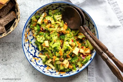 Frischer Brokkoli Salat mit Apfelstücken und Karottenraspeln, serviert in einer blauen Schüssel mit Holzkochlöffeln.
