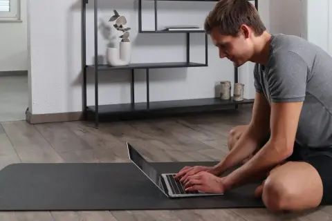 Mann auf Yogamatte tippt auf Laptop, um den Buckel wegtrainieren.