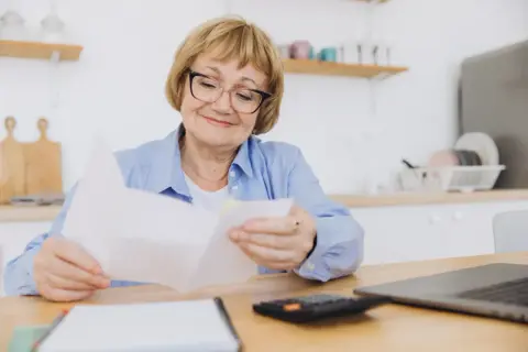 Uśmiechnięta starsza pani analizuje dokumenty, zastanawiając się, o ile wzrośnie emerytura po roku pracy na emeryturze.