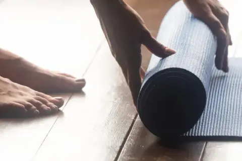 Pieds nus sur un parquet, une main déroule un tapis bleu pour une séance de sport à la maison.