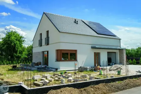 Nowy dom w trakcie budowy, z panelami słonecznymi na dachu. Rozbudowa domu postępuje, widać materiały budowlane i fundamenty.