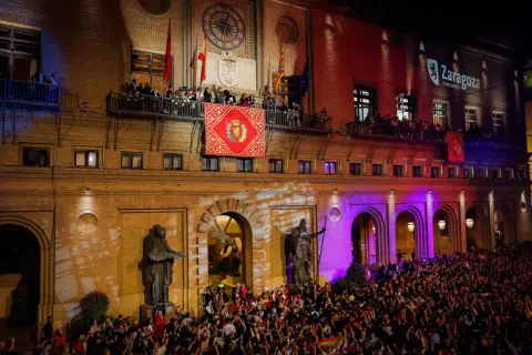 Multitud celebra el pregon de pilares en Zaragoza. Gente en balcones y plaza, luces y banderas.