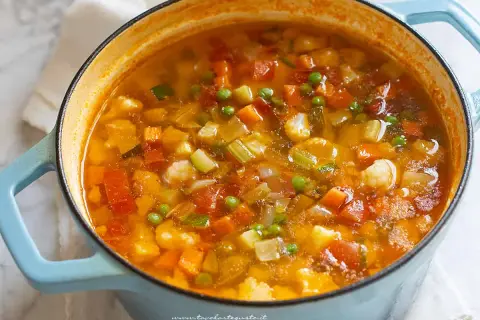Un fumante minestrone di verdure in una casseruola blu, ricco di carote, zucchine, piselli e cavolfiore.