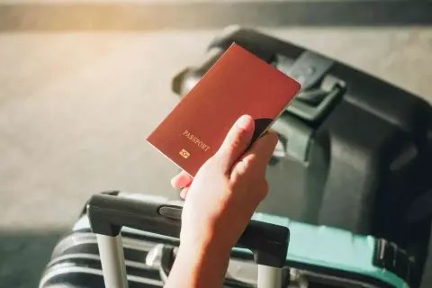 Dłoń trzyma paszport, obok walizka. Problem z wyrabianiem nowego dokumentu z powodu braku starego paszportu.