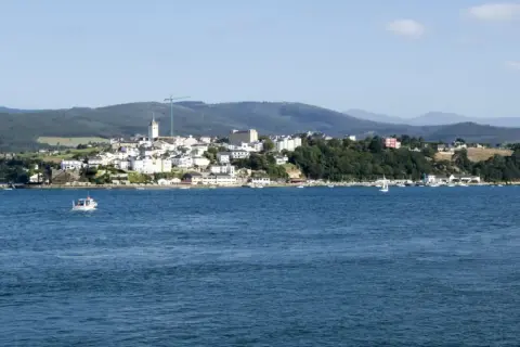 Vista del mirador de Ribadeo con casas blancas y el mar azul. Un pequeño barco navega en la distancia.
