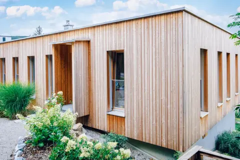 Modernes Haus mit einer Mischfassade aus Holz und Putz. Vertikale Holzverkleidung umrahmt schmale Fenster. Grüne Pflanzen schmücken den Eingangsbereich.