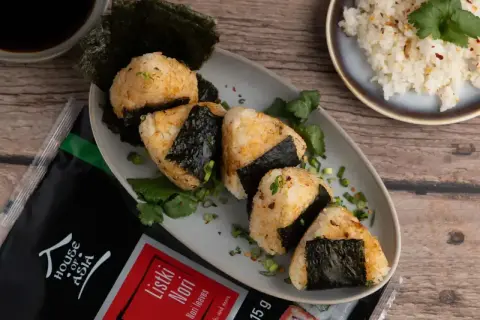Pięć onigiri z ryżem, udekorowanych listkami nori i szczypiorkiem, obok opakowania nori i miseczki z ryżem.