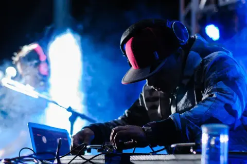Un techno dj con cuffie e cappellino rosso mixa musica su un palco illuminato da luci blu e fumo.