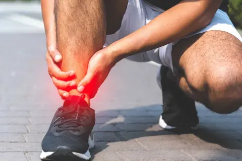 Osoba trzymająca się za bolesną, czerwoną kostkę w bucie sportowym.
