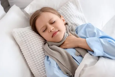 Dziewczynka w niebieskiej piżamie i szaliku trzyma się za bolące gardło, leżąc w łóżku.