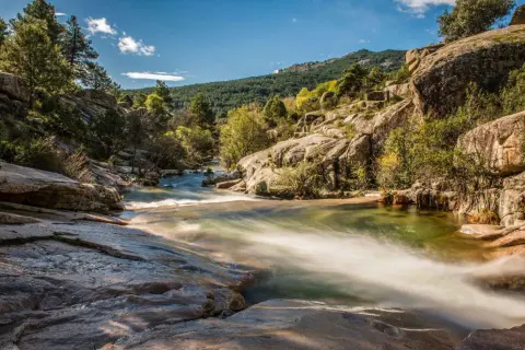 Río serpenteante entre rocas y vegetación, con una montaña boscosa al fondo, un paisaje idílico cerca de Madrid.