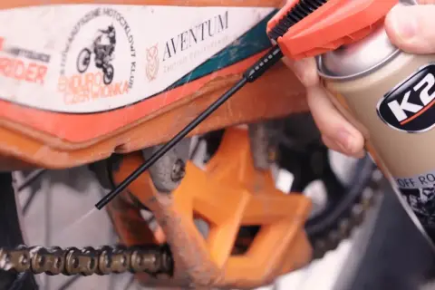 Ręka trzymająca puszkę smaru K2 i aplikująca go na łańcuch motocykla.
