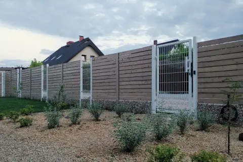 Nowoczesne ogrodzenie z drewnianych desek i metalowych słupków, z furtką. Czy ogrodzenie jest obiektem budowlanym? W tle dom.