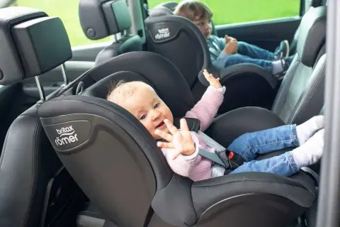 Uśmiechnięte dziecko w foteliku samochodowym. Starsze dziecko z tyłu. Od kiedy można wozić dziecko przodem do kierunku jazdy? Odpowiedź znajdziesz w instrukcji fotelika.