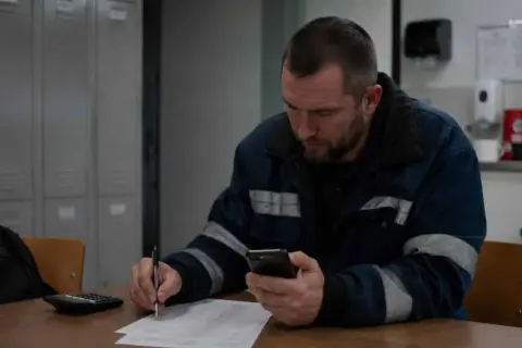 Pracownik w roboczym ubraniu sprawdza telefon, pisząc coś na papierze. Może to być rozliczenie za dodatek za drugą zmianę.