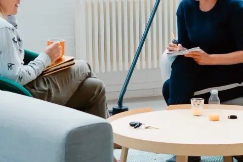 Dwie osoby siedzą w przytulnym pokoju, jedna pije z kubka, druga pisze w notatniku.