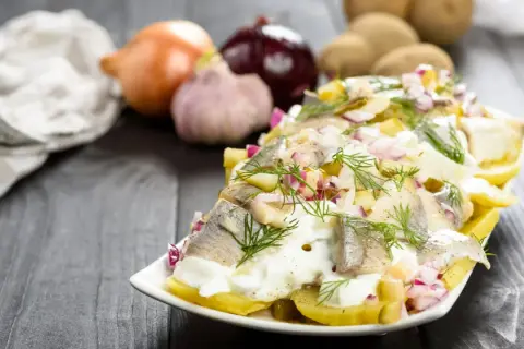 Pyszna sałatka ziemniaczana ze śledziem, udekorowana koperkiem i czerwoną cebulką. Obok leżą ziemniaki, cebula i czosnek.
