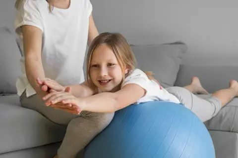 Uśmiechnięta dziewczynka ćwiczy na piłce rehabilitacyjnej, poprawiając swój **tonus mięśniowy** pod okiem mamy.