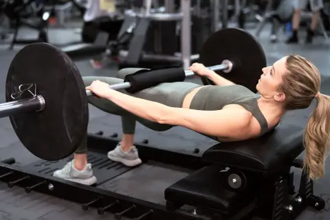 Kobieta wykonuje hip thrust z obciążeniem na ławce. Ćwiczenie to świetnie wzmacnia pośladki i jest możliwe do wykonania nawet w domu.