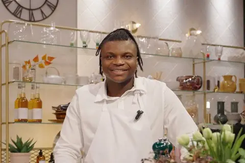 Kelvin Wokomaty, en glad kock i vitt förkläde, ler mot kameran. Bakom honom syns hyllor med flaskor och dekorationer.