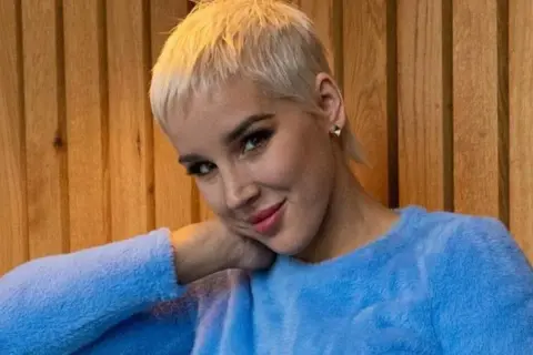 Kobieta z krótkimi blond włosami w niebieskim swetrze opiera głowę na dłoni.